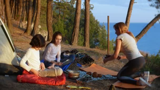 Una familia feliz de turistas, comiendo comida de campamento, junto a una tienda de campaña en el borde de una costa empinada en un pinar con una magnífica vista del paisaje marino. 4k . — Vídeos de Stock