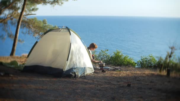 La mujer, descansando, cocina comida junto a la tienda en el borde de una costa empinada en un pinar con una magnífica vista del paisaje marino. 4k . — Vídeos de Stock