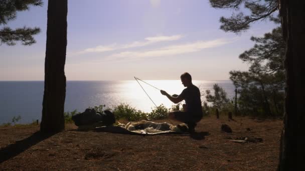 Ein männlicher Tourist richtet ein Zelt am Rande eines steilen Küstenufers in einem Pinienhain mit herrlichem Blick auf das Meer ein. 4k. — Stockvideo