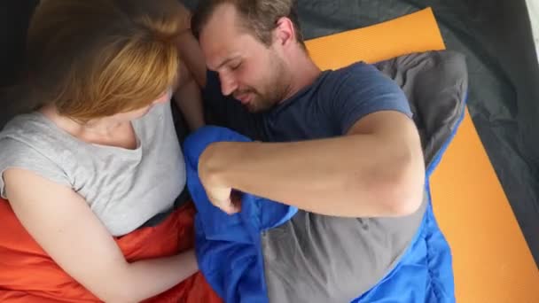 Fecha a porta. Um casal bonito abraçando dentro de uma tenda deitada em sacos de dormir, eles flertam e beijam, 4k . — Vídeo de Stock