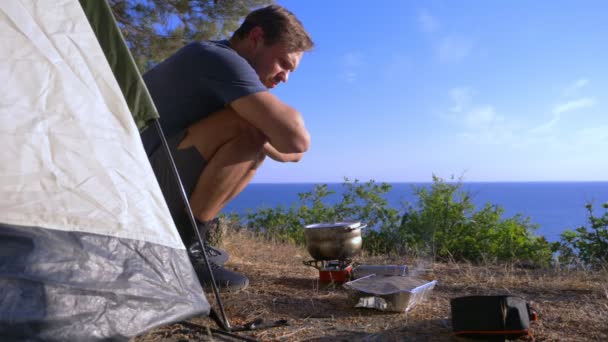 Un hombre los campistas, cocina comida al lado de una tienda de campaña en el borde de una costa empinada en un pinar con una magnífica vista del paisaje marino. 4k — Vídeo de stock