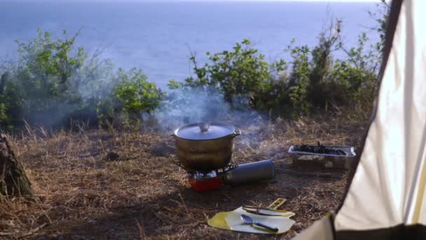 Un bombín y una parrilla desechable. cocinar al lado de una tienda de campaña en el borde de una costa empinada en un pinar con una magnífica vista del paisaje marino. 4k . — Vídeo de stock