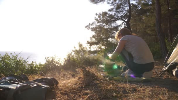 La mujer, descansando, cocina comida junto a la tienda en el borde de una costa empinada en un pinar con una magnífica vista del paisaje marino. 4k . — Vídeos de Stock