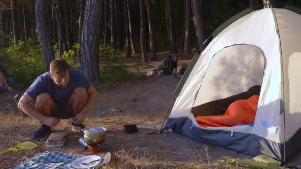 Un hombre los campistas, cocina comida al lado de una tienda de campaña en el borde de una costa empinada en un pinar con una magnífica vista del paisaje marino. 4k — Vídeos de Stock