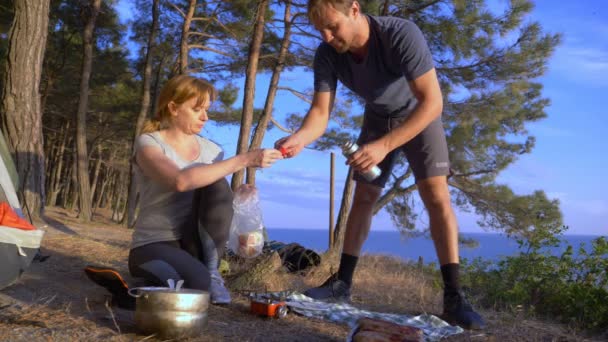 Un hombre y una mujer, los campistas, cocina comida al lado de una tienda de campaña en el borde de una costa empinada en un pinar con una magnífica vista del paisaje marino. 4k — Vídeo de stock