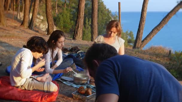 Une famille heureuse de touristes, manger de la nourriture de camp, à côté d'une tente sur le bord d'une côte escarpée dans une pinède avec une vue magnifique sur le paysage marin. 4k . — Video