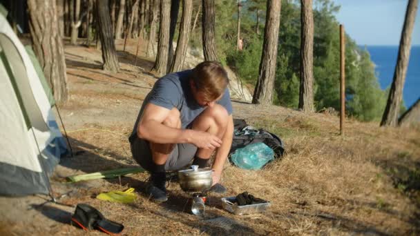 Un hombre los campistas, cocina comida al lado de una tienda de campaña en el borde de una costa empinada en un pinar con una magnífica vista del paisaje marino. 4k — Vídeos de Stock