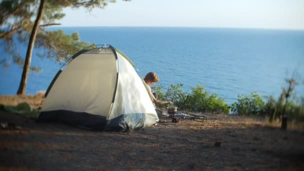 De vrouw, rusten, kookt eten naast de tent aan de rand van een steile kustlijn in een pijnbomenbos met een prachtig uitzicht op het Zeegezicht. 4k. — Stockvideo