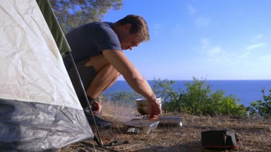 bir adam kampçılar, aşçılar yemek denizin muhteşem manzaralı pine Grove dik bir sahil kenarında bir çadır yanındaki manzara. 4k