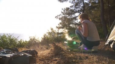 dinlenme, kadın, yanında çadır deniz manzarası muhteşem manzaralı pine Grove dik bir sahil kenarında yemek pişiriyor. 4k.