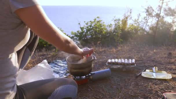 Женщина, отдыхая, готовит еду рядом с палаткой на краю крутой береговой линии в сосновой роще с великолепным видом на море. 4k . — стоковое видео