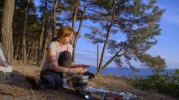 La femme, au repos, cuisine à côté de la tente sur le bord d'une côte escarpée dans une pinède avec une vue magnifique sur le paysage marin. 4k . — Video