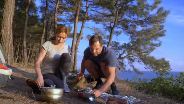 Un hombre y una mujer, los campistas, cocina comida al lado de una tienda de campaña en el borde de una costa empinada en un pinar con una magnífica vista del paisaje marino. 4k — Vídeos de Stock