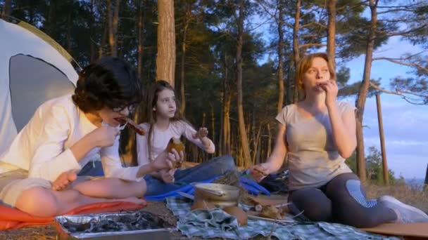 Una familia feliz de turistas, comiendo comida de campamento, junto a una tienda de campaña en el borde de una costa empinada en un pinar con una magnífica vista del paisaje marino. 4k . — Vídeo de stock