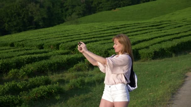 Mulher feliz, um blogueiro faz fotos e vídeos em seu telefone para redes sociais enquanto caminha por uma plantação de chá durante uma excursão. conceito de viagem, lazer. Estilo de vida. 4k . — Vídeo de Stock