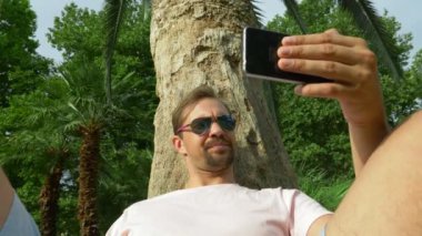 Bir cep telefonu ile bir palmiye ağacının altında oturan genç turist. Olumlu duygular. Zaman rahatla. 4k, ağır çekim