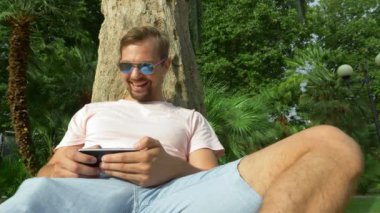 Bir cep telefonu ile bir palmiye ağacının altında oturan genç turist. Olumlu duygular. Zaman rahatla. 4k, ağır çekim.