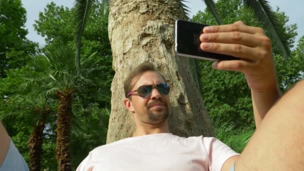 Bir cep telefonu ile bir palmiye ağacının altında oturan genç turist. Olumlu duygular. Zaman rahatla. 4k, ağır çekim — Stok video
