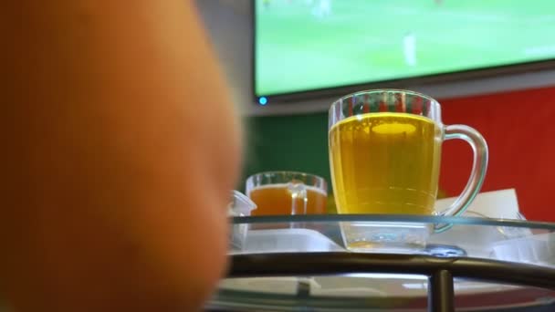 A ver futebol na televisão no pub local. Comida, cerveja. uma caneca com cerveja em foco, uma tela de TV com uma transmissão de um borrão de futebol. 4k — Vídeo de Stock