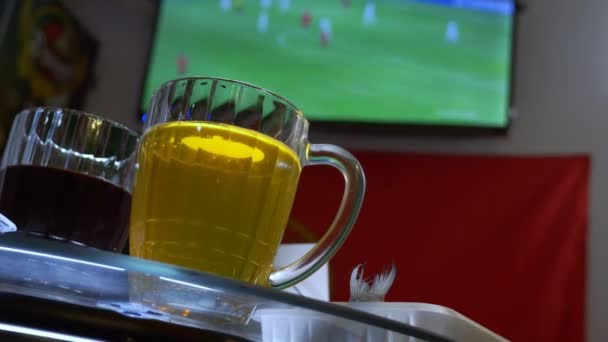 Sledovat fotbalový zápas v televizi v místní hospodě. Jídlo, pivo. hrnek s pivem v zaměření, televizní obrazovka s vysílání fotbalu rozostření. 4k — Stock video