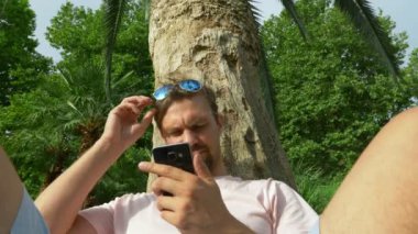 Bir cep telefonu ile bir palmiye ağacının altında oturan genç turist. Olumlu duygular. Zaman rahatla. 4k, ağır çekim.