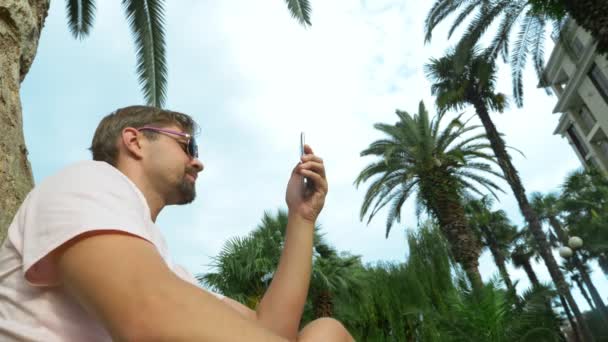 Unga turist sitter under en Palm med en mobiltelefon. Positiva känslor. Tid koppla av. 4k, Slowmotion — Stockvideo