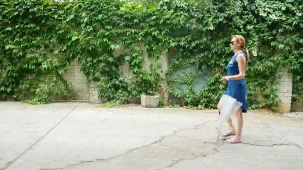 Žena v modrých šatech kráčí podél bílé zdi. Ona drží slunečník a plážová taška 4k — Stock video