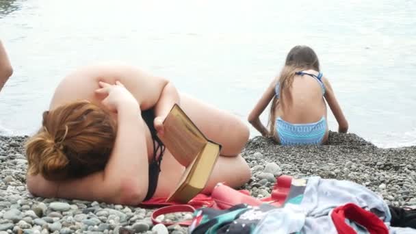 Una mujer gorda irreconocible yace en la playa y lee un libro, en el fondo, la gente delgada se divierte en el mar. 4k — Vídeos de Stock