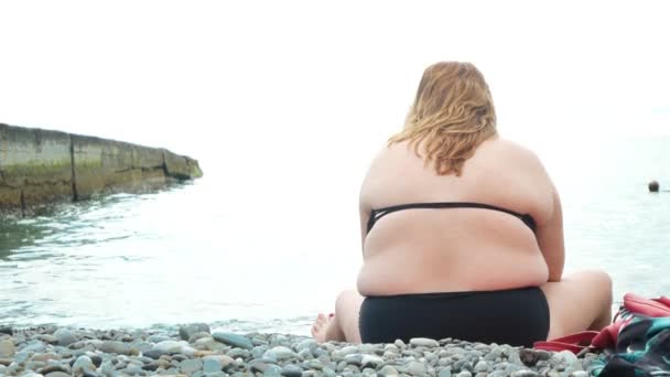 Egy kövér, felismerhetetlen nő ül a parton vele vissza, hogy a kamera, boldog emberek úszni a tengerben. 4k — Stock videók