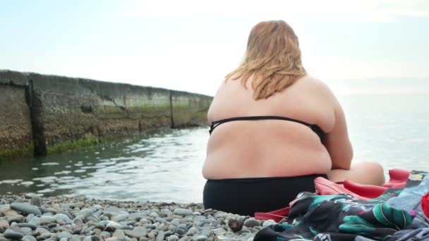 A fat unrecognizable woman is sitting on the beach with her back to the camera, happy people are swimming in the sea. 4k — Stock Video