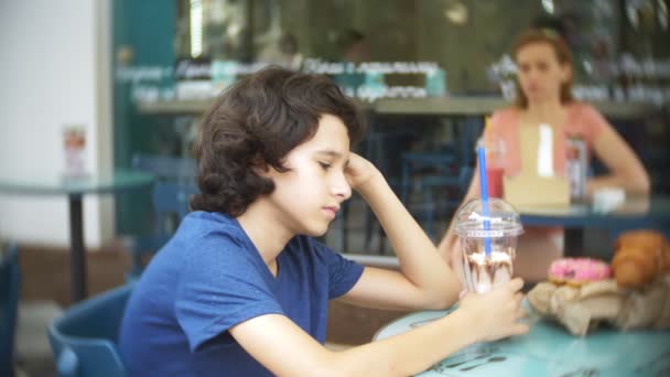 Θλιβερή αγόρι έφηβος κάθεται σε ένα τραπέζι του καφέ, κρατώντας ένα milkshake με σοκολάτα στο χέρι. 4 k. αργή κίνηση. — Αρχείο Βίντεο