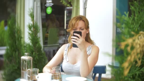 Δυσαρεστημένοι κορίτσι βλέποντας τηλέφωνο κάθεται σε μια καφετέρια στο δρόμο — Αρχείο Βίντεο