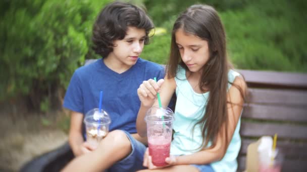 Niño y niña adolescente sentado en un banco, sosteniendo vasos con batidos frescos y en la mano. 4k. Movimiento lento . — Vídeos de Stock