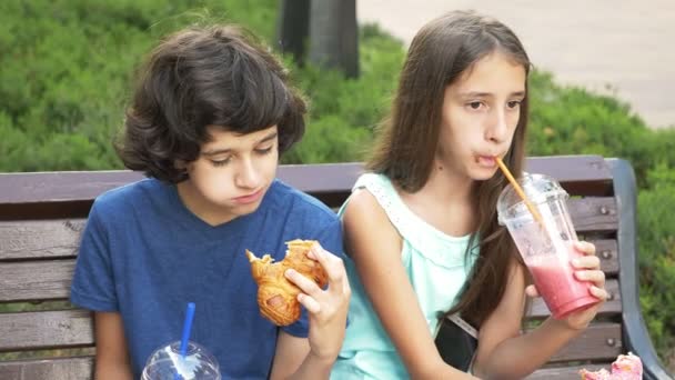 Triste chico y adolescente chica sentado en banco sosteniendo gafas con frescos y batidos en la mano. comer un donut y un croissant de 4k. Movimiento lento . — Vídeos de Stock