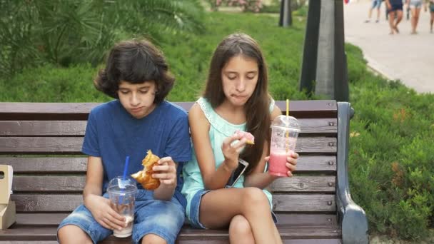 Ein trauriger Junge und ein Teenager sitzen auf einer Bank und halten Gläser mit frischen Smoothies in der Hand. Essen Sie einen Donut und ein 4k Croissant. Zeitlupe. — Stockvideo