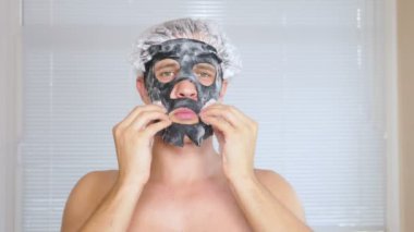 Yüz paketi olan garip adam. Genç bir adam bir ev bir ayna önünde yüz için kozmetik işlemler yapar. 4k