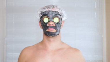 Yüz paketi olan garip adam. Genç bir adam bir ev bir ayna önünde yüz için kozmetik işlemler yapar. 4k, salatalık maskesi