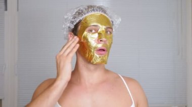 Yüz paketi olan garip adam. Genç bir adam bir ev bir ayna önünde yüz için kozmetik işlemler yapar. 4k, altın maske