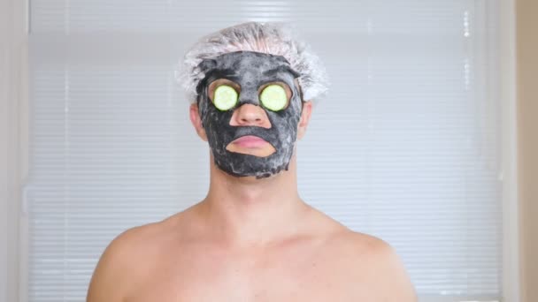 Странный человек с лицом. Молодой человек делает косметические процедуры для лица дома перед зеркалом. 4k, огуречная маска — стоковое видео