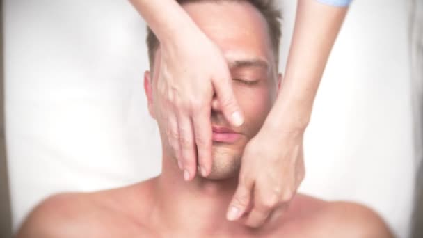 Un hombre recibiendo un masaje facial. Masajea la cara y el cuello. tumbado en la oficina del kasmetólogo. 4k, cámara lenta, vista superior — Vídeos de Stock