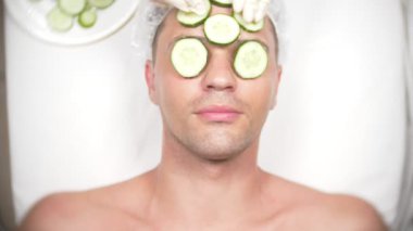 Yüz paketi olan garip adam. Genç bir adam bir ev bir ayna önünde yüz için kozmetik işlemler yapar. 4k, salatalık maskesi