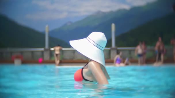 Медленное движение, закрытие, портрет. молодая женщина в большой белой шляпе, загорает и отдыхает в солнечный день в роскошном бассейне на фоне горного пейзажа. горный курорт с открытым бассейном. 4k — стоковое видео