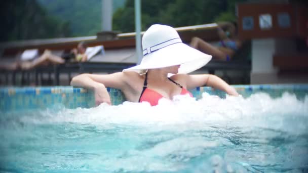 Lento movimento, fecho, reboque. jovem mulher em um chapéu branco grande, banhar-se de sol e relaxar em um dia ensolarado em uma piscina de luxo em um fundo de uma paisagem montesa. resort de montanha com piscina exterior. 4k — Vídeo de Stock