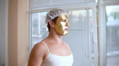Yüz paketi olan garip adam. Genç bir adam bir ev bir ayna önünde yüz için kozmetik işlemler yapar. 4k, altın maske