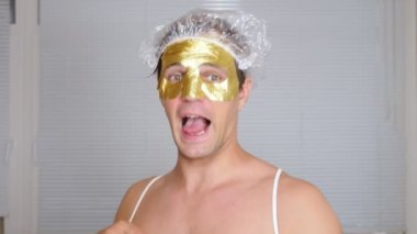 Yüz paketi olan garip adam. Genç bir adam bir ev bir ayna önünde yüz için kozmetik işlemler yapar. 4k, altın maske