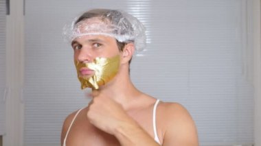 Yüz paketi olan garip adam. Genç bir adam bir ev bir ayna önünde yüz için kozmetik işlemler yapar. 4k, altın maske