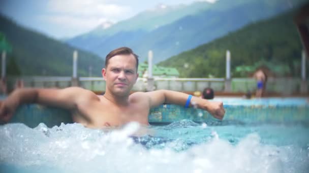Lassú mozgás, szoros, portré. a fiatalember napozhat, és pihenjen egy napsütéses napon a háttérben egy hegyi táj a fényűző úszómedence állnak rendelkezésre. Mountain resort szabadtéri medencével rendelkezik. 4k — Stock videók