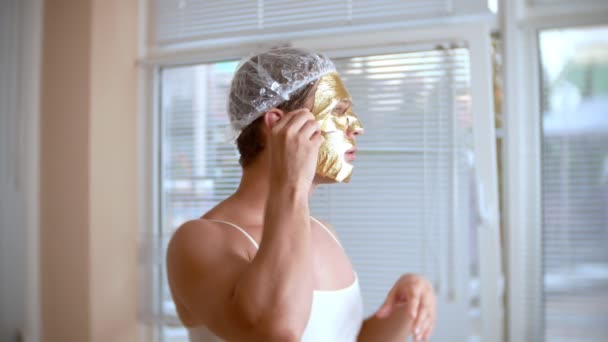 Seltsamer Mann mit Gesichtsmaske. Ein junger Mann macht kosmetische Eingriffe am Gesicht eines Hauses vor dem Spiegel. 4k, goldene Maske — Stockvideo