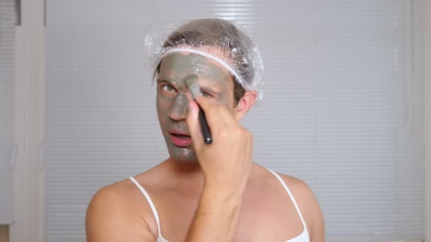 Vreemde man met gezicht pack. Een jonge man doet cosmetische ingrepen voor het gezicht van een huis voor een spiegel. 4k — Stockvideo