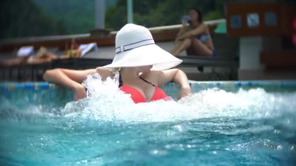 Lento movimento, fecho, reboque. jovem mulher em um chapéu branco grande, banhar-se de sol e relaxar em um dia ensolarado em uma piscina de luxo em um fundo de uma paisagem montesa. resort de montanha com piscina exterior. 4k — Vídeo de Stock